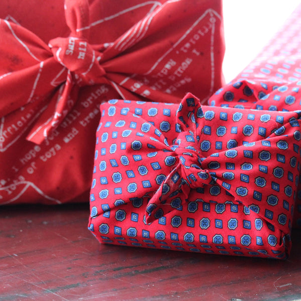 Three gifts wrapped in printed red fabric, tied in Furoshiki style.  The gifts are arranged on a bench on edge with their bows facing forward.
