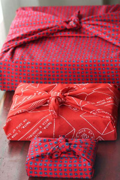 Three gifts wrapped in coordinating patterned red fabric, tied Furoshiki style.  The gifts are lined up on a bench from smallest to largest.