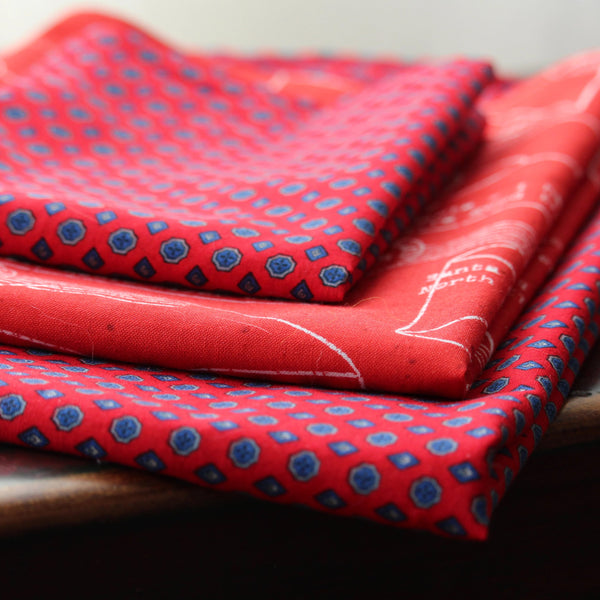 Three squares of fabric gift wrap are folded and stacked on a bench.  The squares are coordinated fabrics that are mostly red with blue dots or white writing featuring letters to Santa.