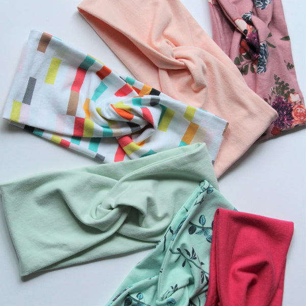 A collection of six jersey knit headbands in spring colours and patterns are fanned out on a white background