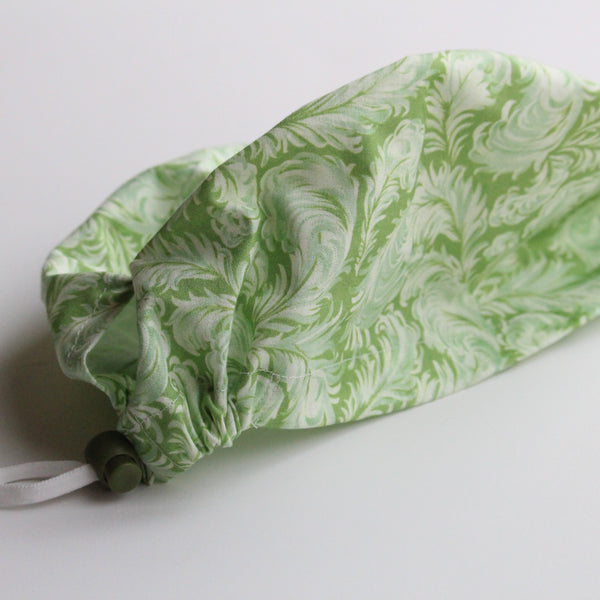 A handmade scrub hat in green with a white feather pattern and a green toggle adjuster lies on a white background.
