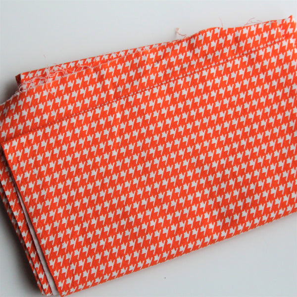 A folded piece of orange and white houndstooth cotton fabric lies on a white background.