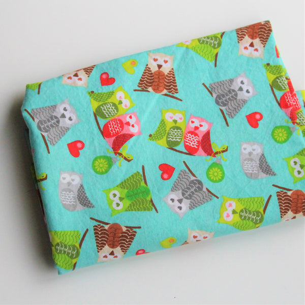 A folded piece of turquoise fabric with lime green, grey, brown and red owls with heart shaped eyes lies on a white background.